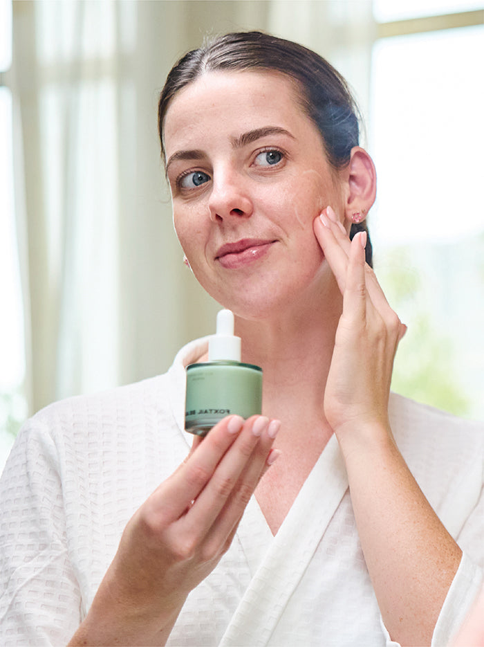 Woman applying Foxtail Beauty Serum to her face with a dropper bottle and jar in a bright room.
