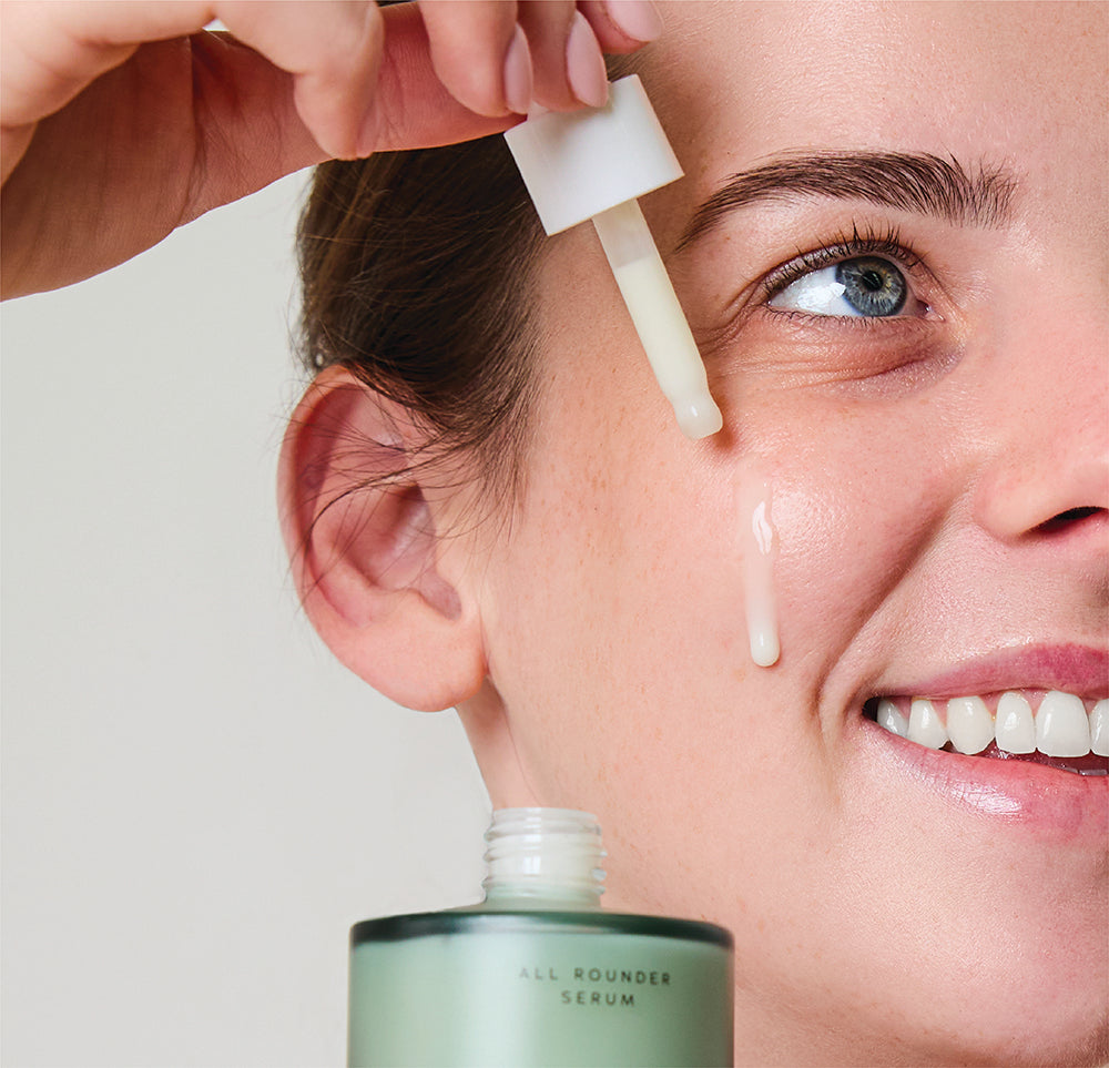 Woman applying a brightening serum for uneven skin tone to her face with a dropper, next to a bottle labeled 'Foxtail Beauty All Rounder Serum'.