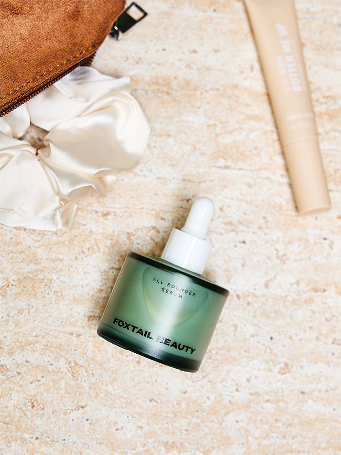 Foxtail Beauty serum bottle on a textured surface with a brown bag and white flowers.